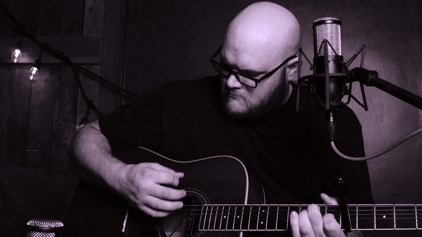 John, in glasses with a beard, playing an acoustic guitar into a studio microphone in a dimly lit room.